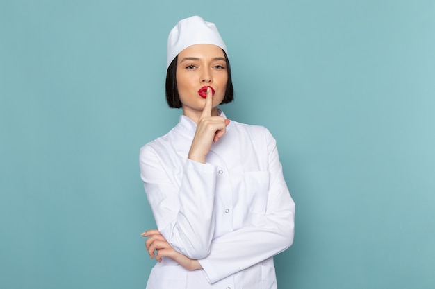 Uma jovem enfermeira com terno branco posando e mostrando o sinal de silêncio na mesa azul. Médico do hospital.