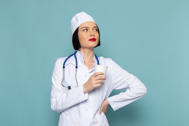 Uma jovem enfermeira com vista frontal, vestida com um terno médico branco e um estetoscópio azul, segurando um copo de plástico na mesa azul.