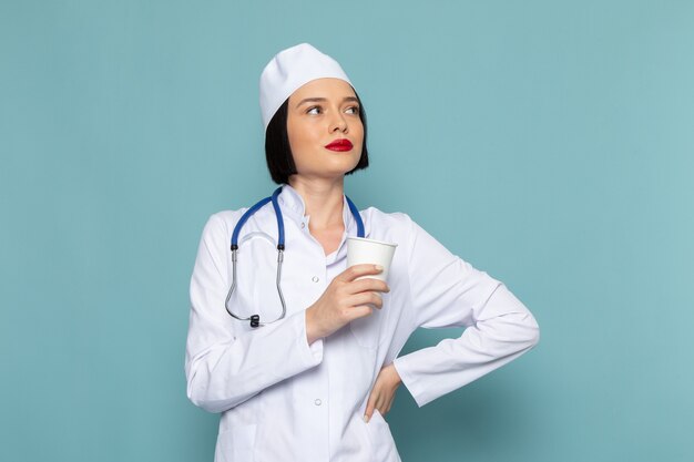 Uma jovem enfermeira com vista frontal, vestida com um terno médico branco e um estetoscópio azul, segurando um copo de plástico na mesa azul.