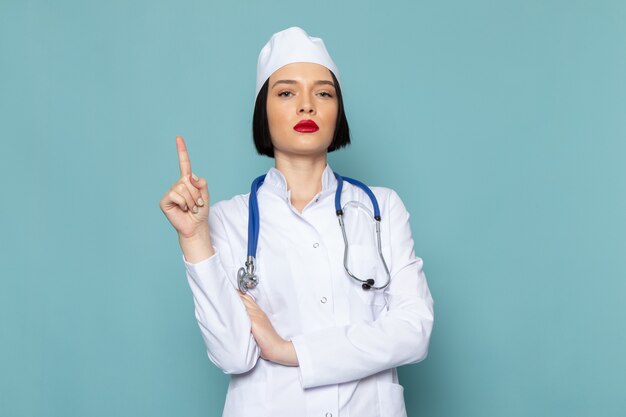 Uma jovem enfermeira de vista frontal em um terno médico branco e um estetoscópio azul posando com o dedo levantado na mesa azul.