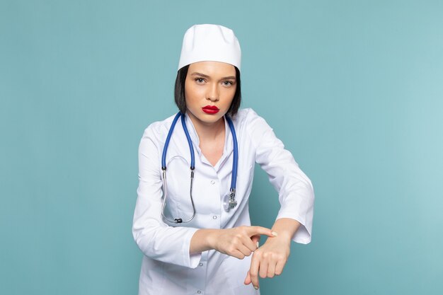 Uma jovem enfermeira de vista frontal em um terno médico branco e um estetoscópio azul posando sobre a mesa azul.