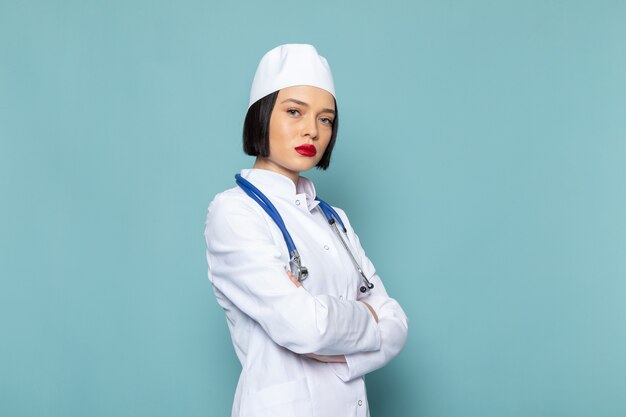 Uma jovem enfermeira de vista frontal em um terno médico branco e um estetoscópio azul posando sobre a mesa azul.