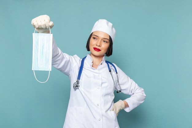 Uma jovem enfermeira, vista de frente, em um terno médico branco e luvas brancas com estetoscópio azul, segurando uma máscara na mesa azul.