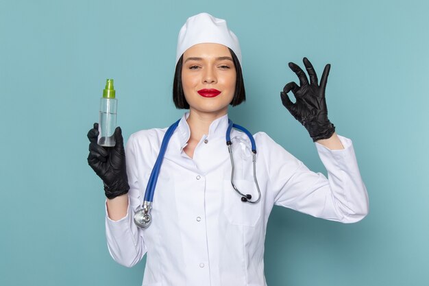 Uma jovem enfermeira, vista frontal, em um terno médico branco e um estetoscópio azul segurando spray na mesa azul