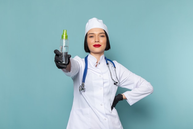 Uma jovem enfermeira, vista frontal, em um terno médico branco e um estetoscópio azul, segurando um frasco de spray na mesa azul.