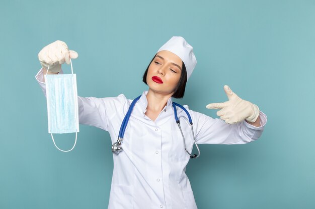 Uma jovem enfermeira, vista frontal, em um terno médico branco e um estetoscópio azul, segurando uma máscara branca na mesa azul.