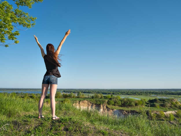 Uma jovem está de pé com os braços erguidos para o céu. Mulher jovem relaxada olhando para a vista. Menina pacífica de pé perto de um penhasco, apreciando a paisagem. - ao ar livre. Retrato de corpo inteiro