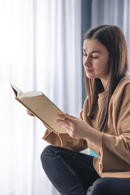 Uma jovem está lendo um livro enquanto está sentada em uma poltrona perto da janela