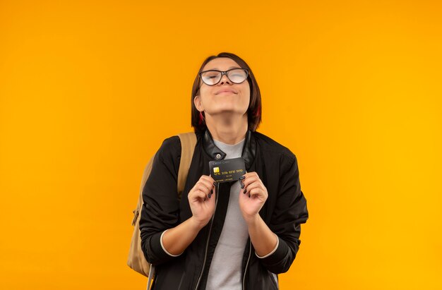 Uma jovem estudante satisfeita usando óculos e uma bolsa com as costas segurando um cartão de crédito com os olhos fechados, isolado na parede laranja