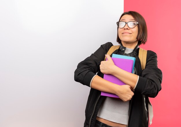 Uma jovem estudante satisfeita usando óculos e uma bolsa traseira segurando livros em frente a uma parede branca isolada na parede rosa