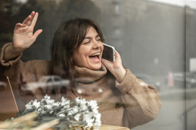 Uma jovem fala ao telefone e acena para alguém, vista da rua.