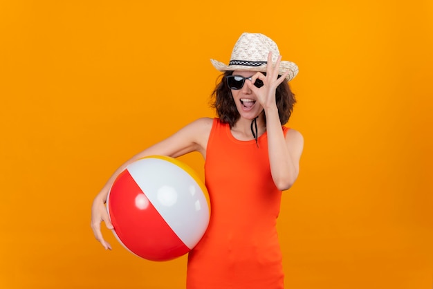 Uma jovem feliz com cabelo curto em uma camisa laranja usando chapéu de sol e óculos escuros segurando uma bola inflável olhando através de um buraco feito com os dedos