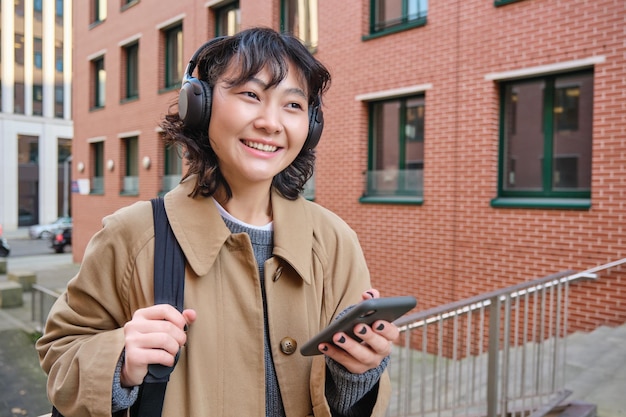 Uma jovem feliz fica na rua com mochila e smartphone ouve música em fones de ouvido espera então