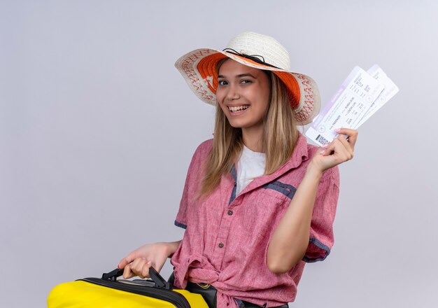 Uma jovem feliz, vestindo uma camisa vermelha, segurando uma mala amarela e mostrando passagens de avião enquanto olha para uma parede branca