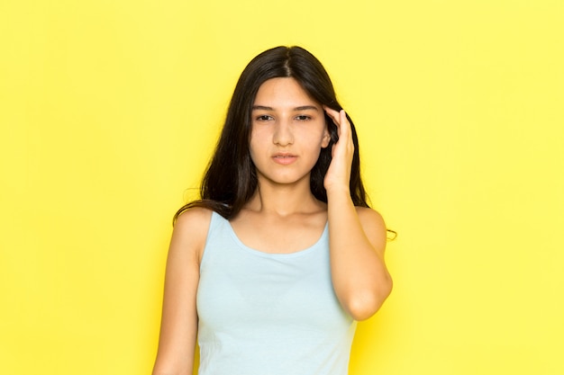 Uma jovem fêmea de camisa azul com uma dor de cabeça de frente no fundo amarelo.