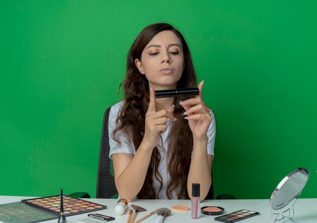 Uma jovem garota bonita sentada à mesa de maquiagem com ferramentas de maquiagem segurando e olhando para o rímel isolado no fundo verde