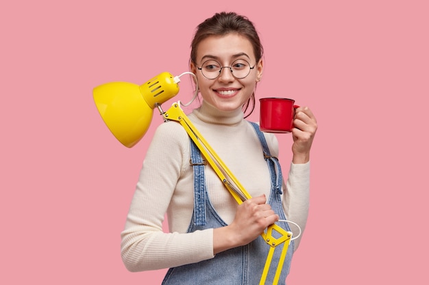 Uma jovem jornalista sorridente trabalha em casa, carrega um abajur amarelo e uma caneca de bebida, parece feliz, faz uma pausa para o café