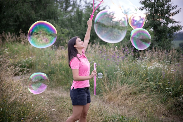Uma jovem lança grandes bolhas de sabão coloridas entre a grama da natureza.