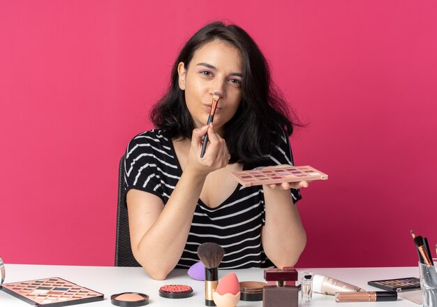 Uma jovem linda e satisfeita se senta à mesa com ferramentas de maquiagem, aplicando a sombra com pincel de maquiagem isolado na parede rosa