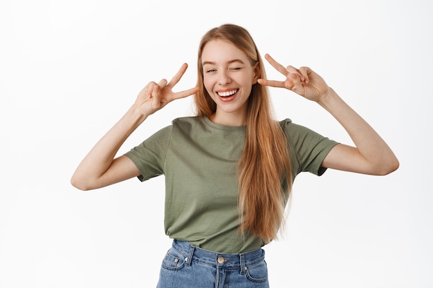 Uma jovem loira positiva e feliz, piscando e sorrindo com os dentes brancos, mostrando o gesto do sinal V da paz perto dos olhos, em pé alegre contra a parede branca