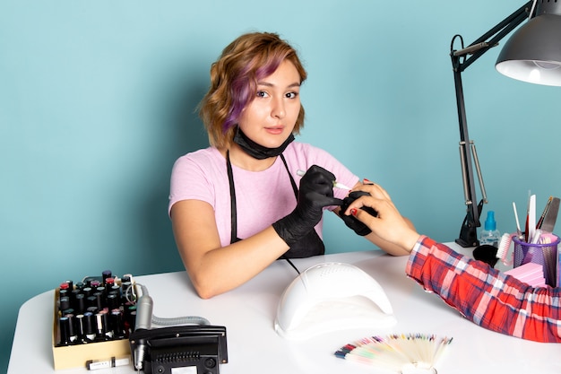 Uma jovem manicure feminina de vista frontal com luvas pretas e máscara preta fazendo manicure em azul