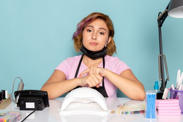 Foto grátis uma jovem manicure feminina de vista frontal com uma camiseta rosa com luvas pretas e uma máscara preta sentada em frente à mesa com uma expressão entediada no azul