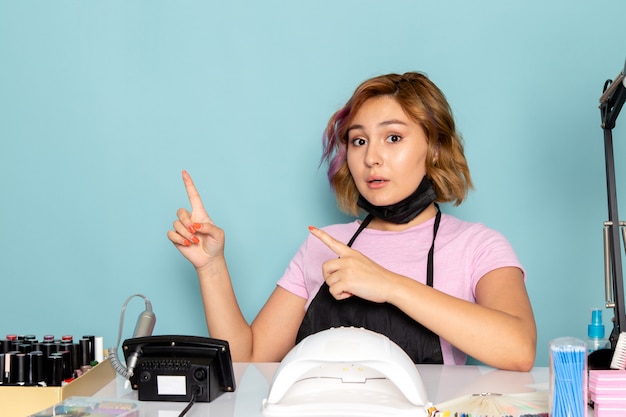 Uma jovem manicure feminina, vista frontal, com uma camiseta rosa com luvas pretas e máscara preta, sentada em frente à mesa apontando para o azul