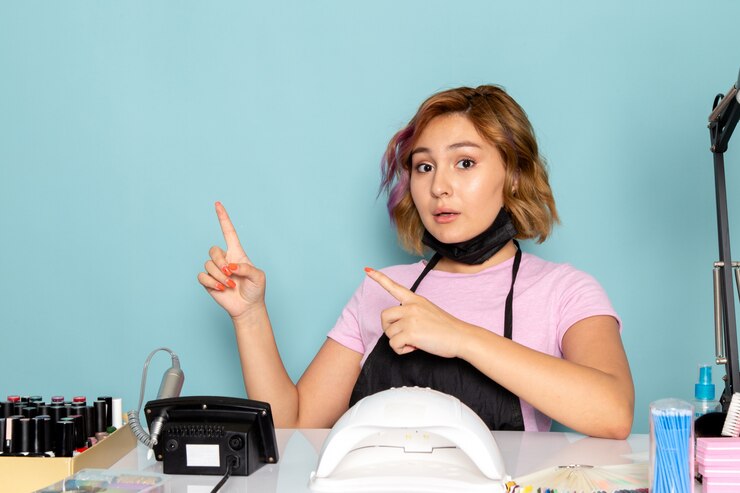 Foto grátis uma jovem manicure feminina, vista frontal, com uma camiseta rosa com luvas pretas e máscara preta, sentada em frente à mesa apontando para o azul