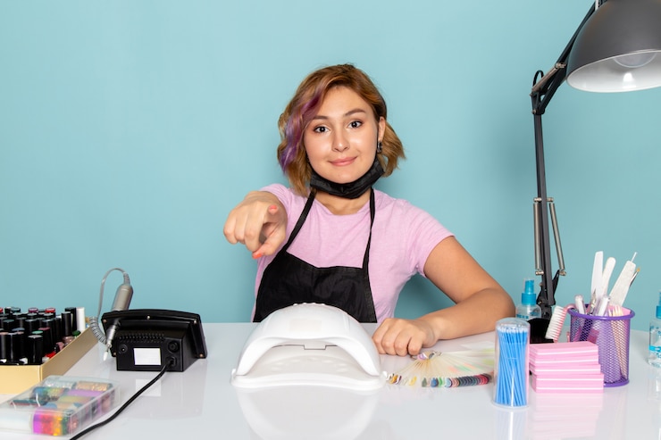 Foto grátis uma jovem manicure feminina, vista frontal, com uma camiseta rosa com luvas pretas e máscara preta sentada em frente à mesa e sorrindo apontando para o azul