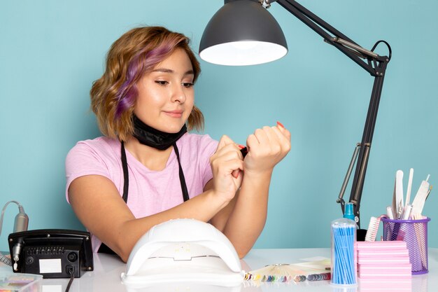 Uma jovem manicure feminina, vista frontal, com uma camiseta rosa com luvas pretas e máscara preta, sentada em frente à mesa, trabalhando com as unhas no azul