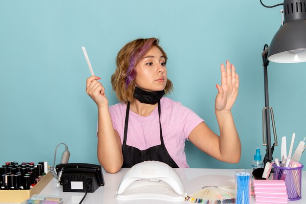 Uma jovem manicure feminina, vista frontal, com uma camiseta rosa com luvas pretas e máscara preta, sentada em frente à mesa, trabalhando com as unhas no azul