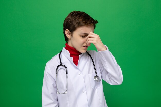 Uma jovem médica de jaleco branco com estetoscópio ao redor do pescoço parecendo indisposta, cansada e trabalhando, tocando o nariz entre os olhos fechados em pé no verde