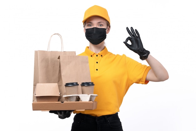 Uma jovem mensageira com uniforme amarelo, luvas pretas e máscara preta, segurando copos de café para entrega de comida