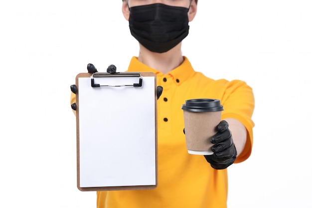 Uma jovem mensageira com uniforme amarelo, luvas pretas e máscara preta, segurando uma xícara de café e um bloco de notas