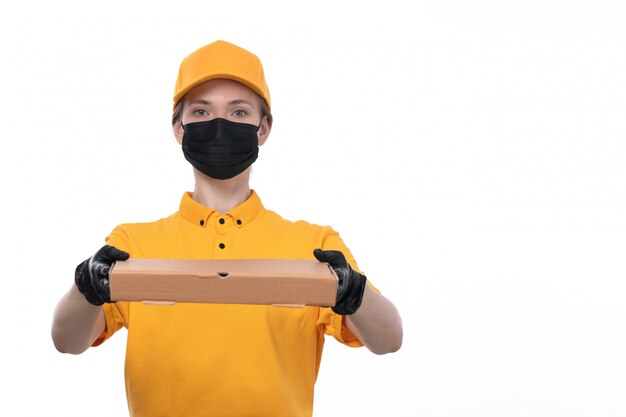 Uma jovem mensageira de frente com luvas pretas de uniforme amarelo e máscara preta segurando uma caixa de comida