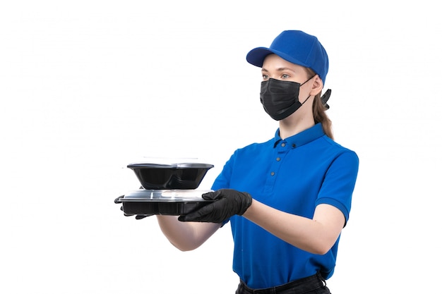 Uma jovem mensageira de frente com luvas pretas de uniforme azul e máscara preta segurando tigelas de entrega de comida