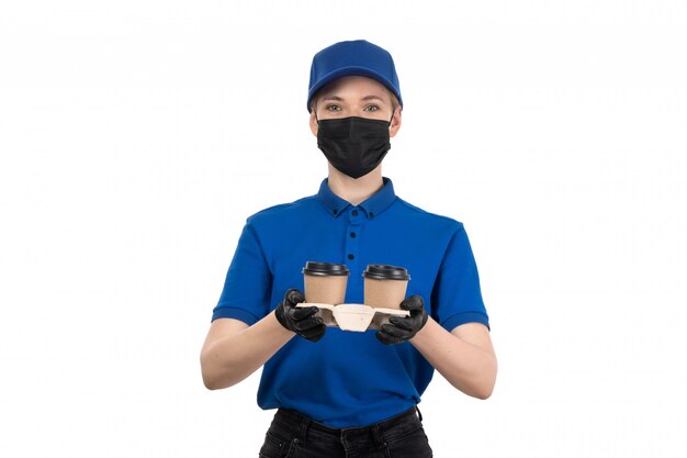 Uma jovem mensageira de frente com uma máscara preta de uniforme azul e luvas segurando xícaras de café