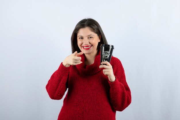 Uma jovem modelo com um suéter vermelho com uma xícara de café apontando para a câmera