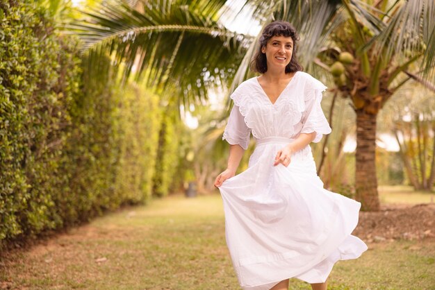 Uma jovem morena caucasiana sorridente usando vestido branco caminha no jardim tropical sozinha na manhã de verão