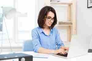 Foto grátis uma jovem morena está digitando no laptop na mesa no escritório. ela usa camisa azul e óculos pretos. ela parece satisfeita com seu trabalho.