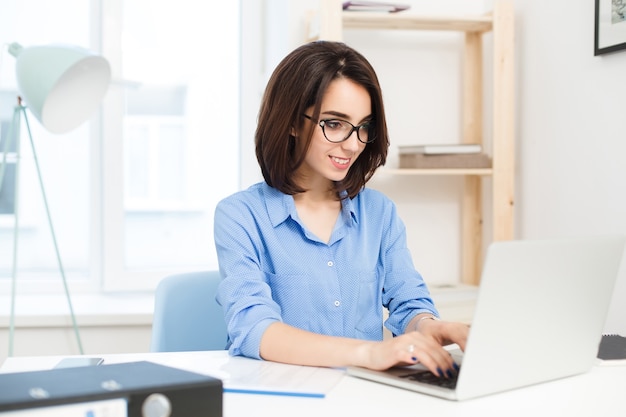 Uma jovem morena está digitando no laptop na mesa no escritório. Ela usa camisa azul e óculos pretos. Ela parece satisfeita com seu trabalho.