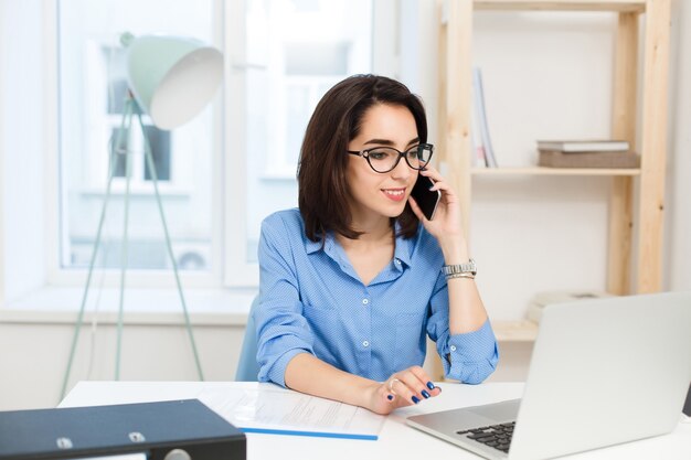 Uma jovem morena está trabalhando na mesa no escritório. Ela usa camisa azul e óculos pretos. Ela está falando ao telefone.