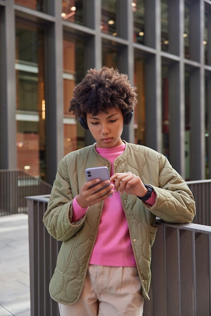 Foto grátis uma jovem muito séria com cabelos cacheados lê mensagem de renda no smartphone moderno fica contra o fundo urbano ouve música via fones de ouvido pesquisa localização via aplicativo de telefone