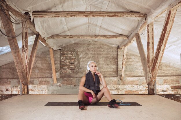 Foto grátis uma jovem mulher atlética em fones de ouvido brancos malhando ouvindo música em uma construção abandonada. descansando após o treinamento. conceito de estilo de vida saudável, esporte, atividade, perda de peso.