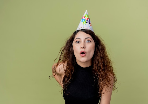 Uma jovem mulher bonita com cabelo encaracolado e chapéu de Natal surpreendeu o conceito de festa de aniversário em pé sobre a parede de luz