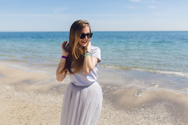 Uma jovem mulher bonita com uma saia cinza exuberante e uma camiseta está em pé perto do mar