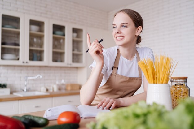 Uma jovem mulher bonita na cozinha, de avental, escreve suas receitas favoritas ao lado de legumes frescos