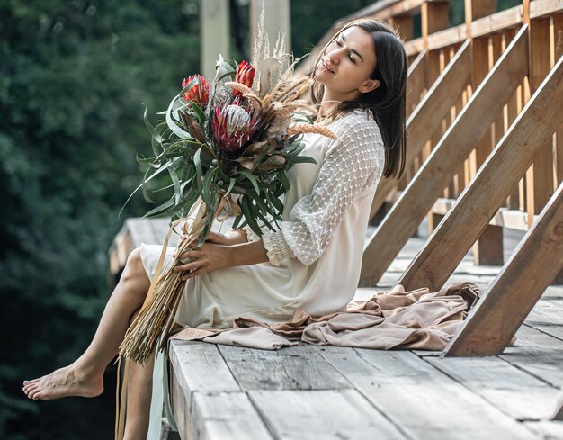 Uma jovem mulher com um vestido branco está sentada em uma ponte de madeira com um buquê de flores exóticas.