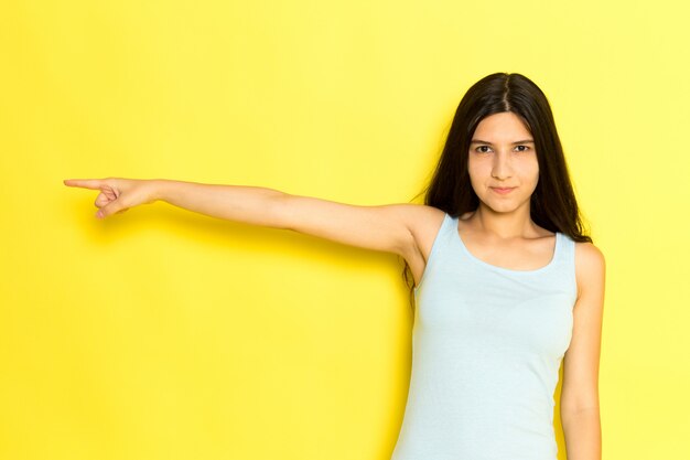 Uma jovem mulher de camisa azul posando e apontando o dedo com expressão de raiva no fundo amarelo.