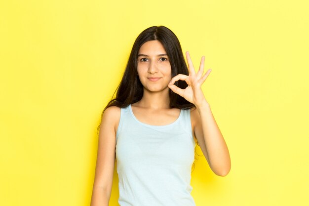 Uma jovem mulher de camisa azul, vista frontal, posando sorrindo e mostrando um sinal de bem